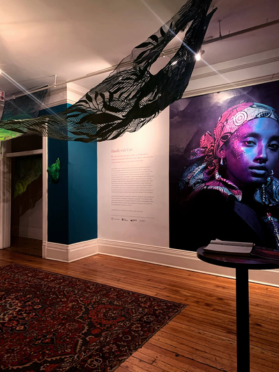 Installation view of large vinyl portrait photograph on wall next to exhibition title and didactics, pictured with hanging artwork depicting plant leaves and foliage made from painted wire mesh.